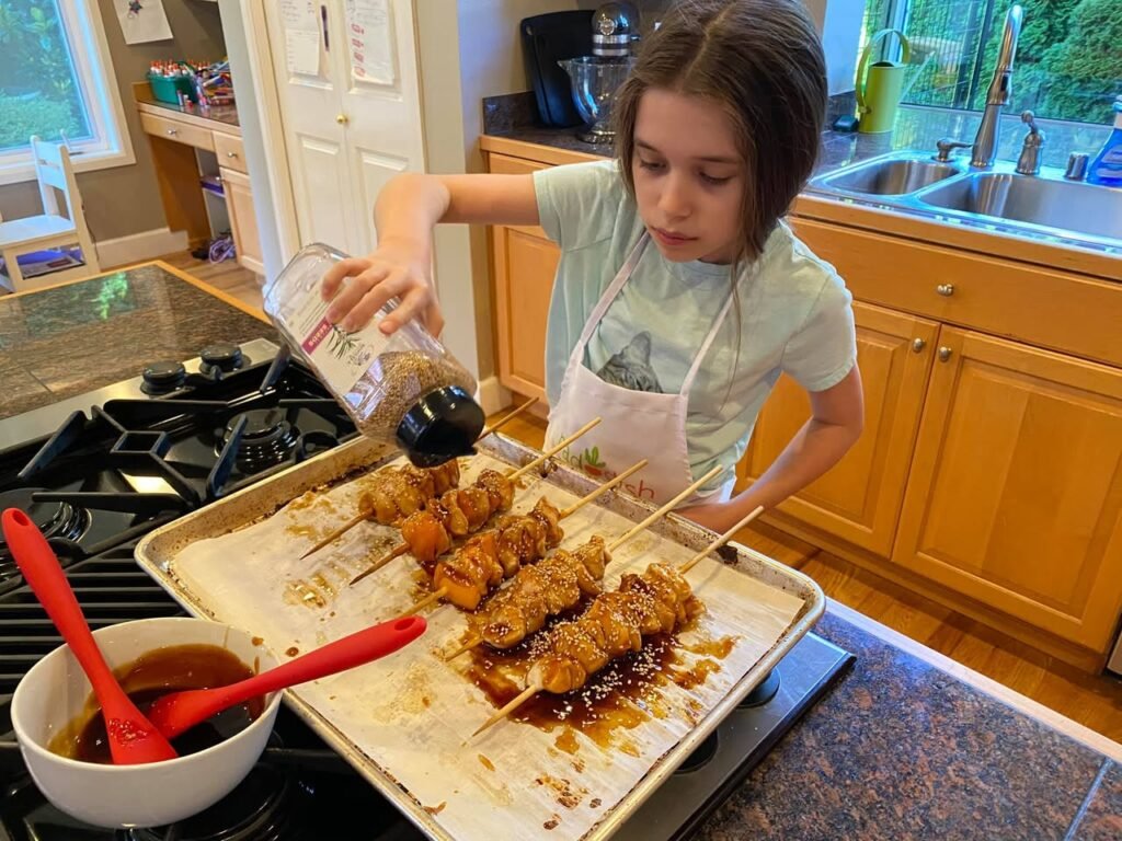 A young person with an apron pouring sauce over skewers of dumplings in a bright kitchen.