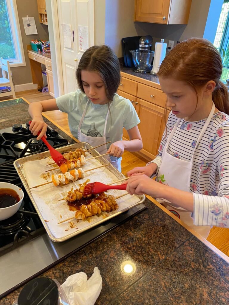 Two children using red brushes to glaze meat rolls on a baking sheet in a home kitchen.