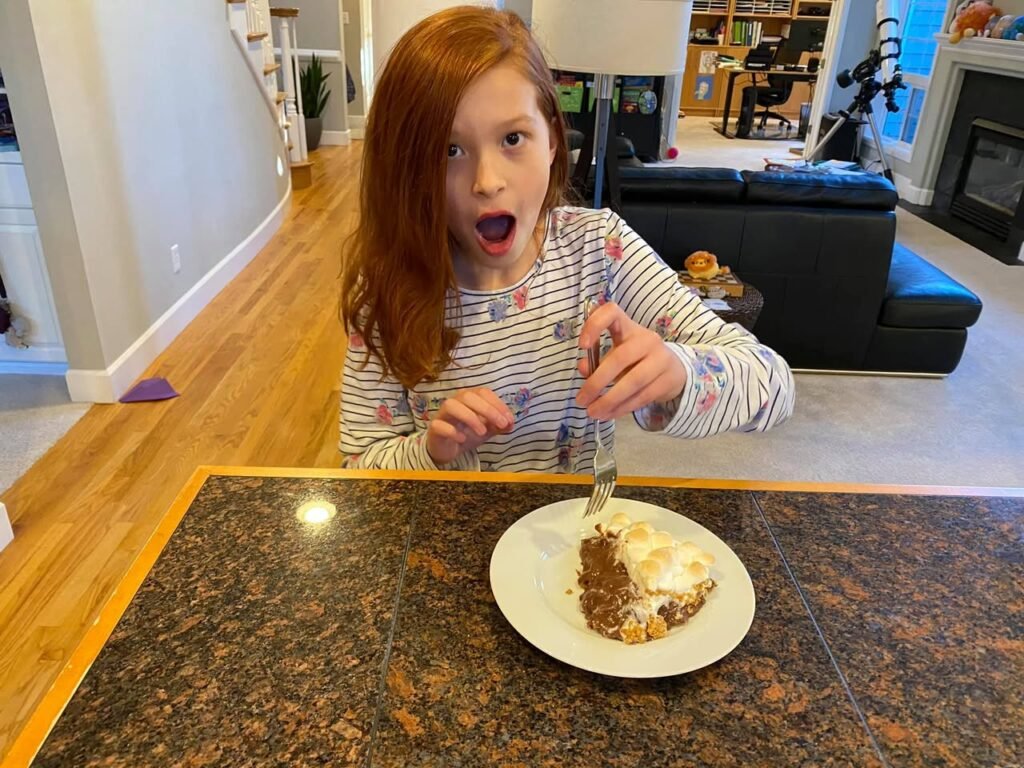 A child with red hair is eating a plate of tacos at a dining table in a bright kitchen.