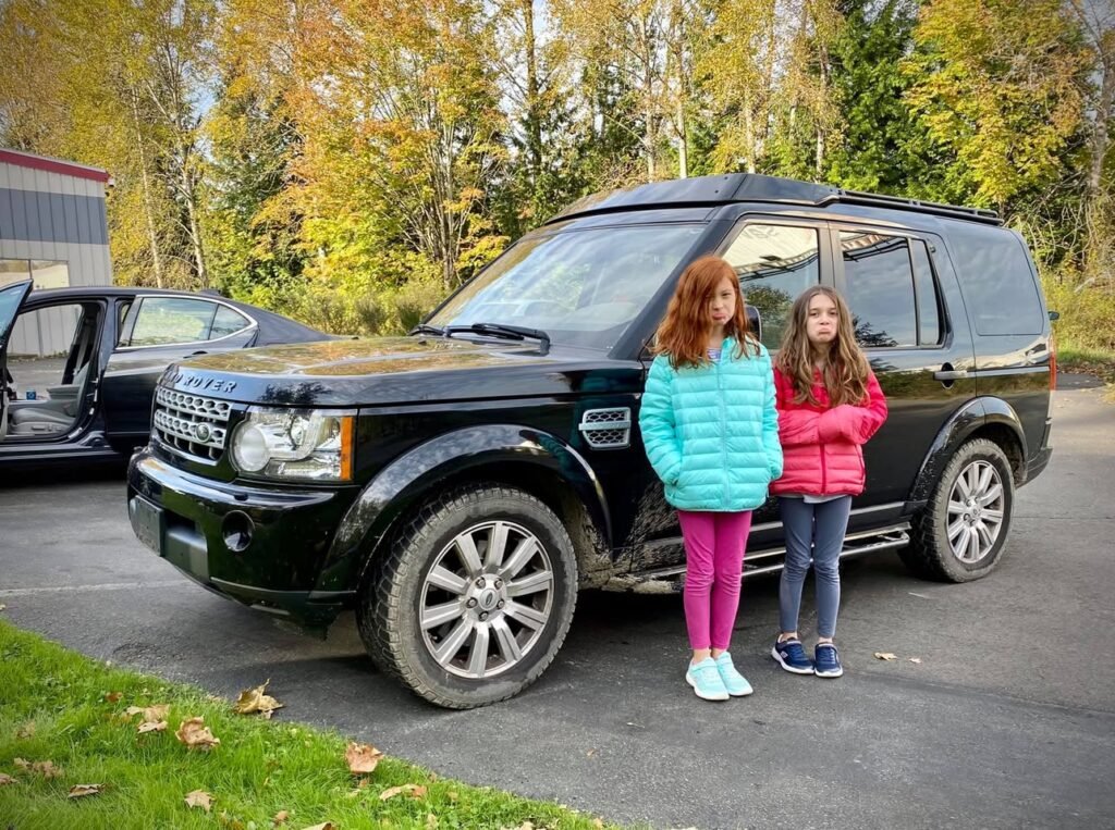 A woman in a bright puffer jacket poses next to a black SUV. The setting features autumn foliage in the background.