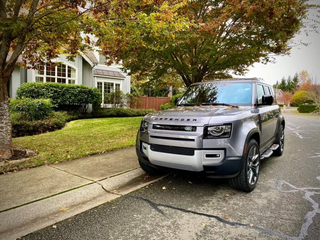A silver Land Rover Defender is parked on a quiet street with a house and autumn trees in the background.