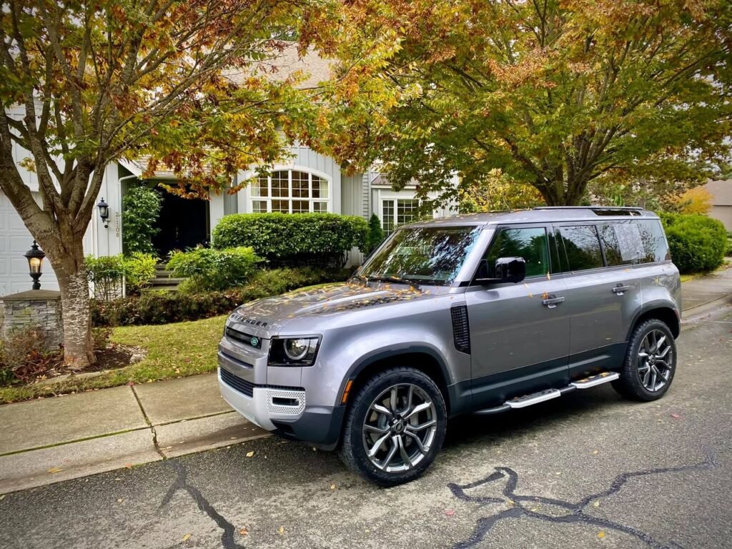 A silver SUV is parked on a quiet street in front of a home with lush foliage.
