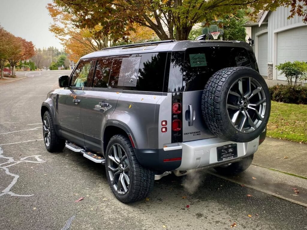 A gray SUV with a spare tire mounted on the rear is parked on a quiet suburban street lined with colorful autumn trees.