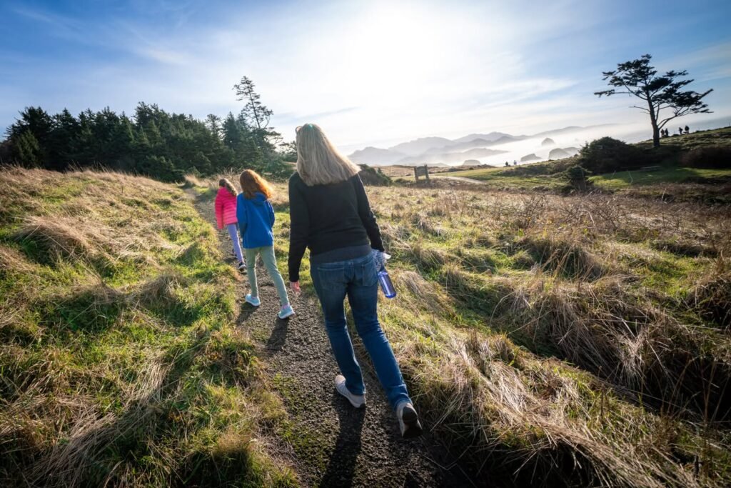 Three people walking along a dirt trail on a grassy hillside with distant mountains and a bright sky. A lone tree clings to the hilltop in the background.