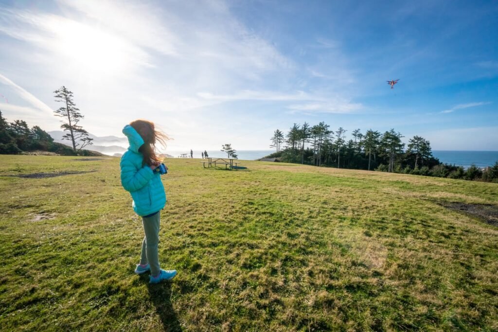 A child in a teal jacket flies a kite in a grassy field near the ocean under a bright blue sky in the Pacific Northwest.