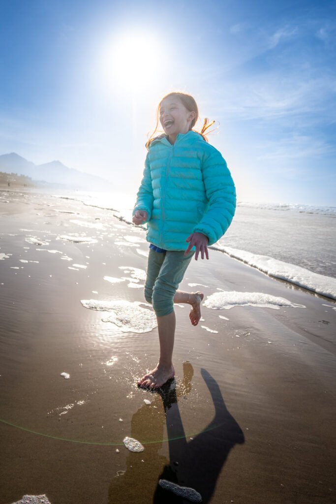 A child in blue walks along a sunny beach, with gentle waves lapping at the shore.
