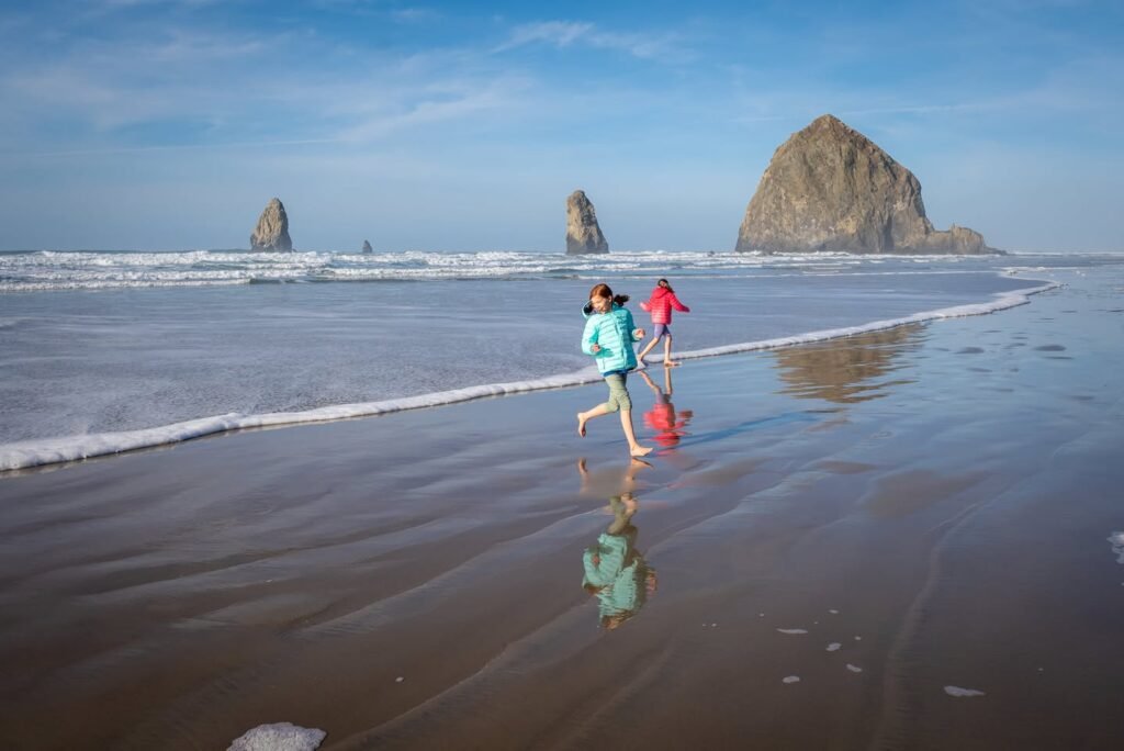 Two people walking on the wet sand at Cannon Beach with sea stacks in the distance under a blue sky.