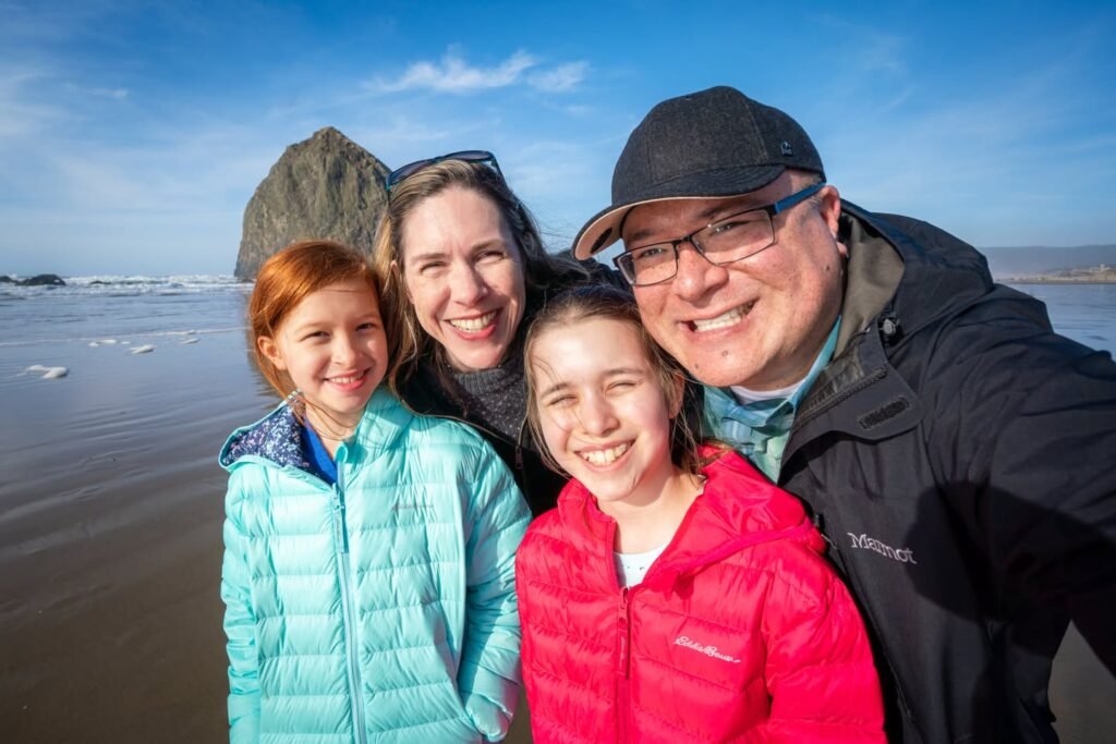 A group of people posing for a photo on a beach with a clear blue sky. One person is in the foreground, while others stand behind, with a rocky hills visible in the distance at Cannon Beach.