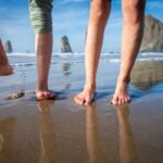 Ecola State Park Barefoot individuals walking on a reflective wet sandy beach with rocky headlands and ocean waves in the distance under a blue sky.