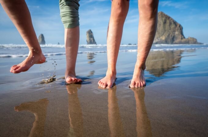 Barefoot individuals walking on a reflective wet sandy beach with rocky headlands and ocean waves in the distance under a blue sky.
