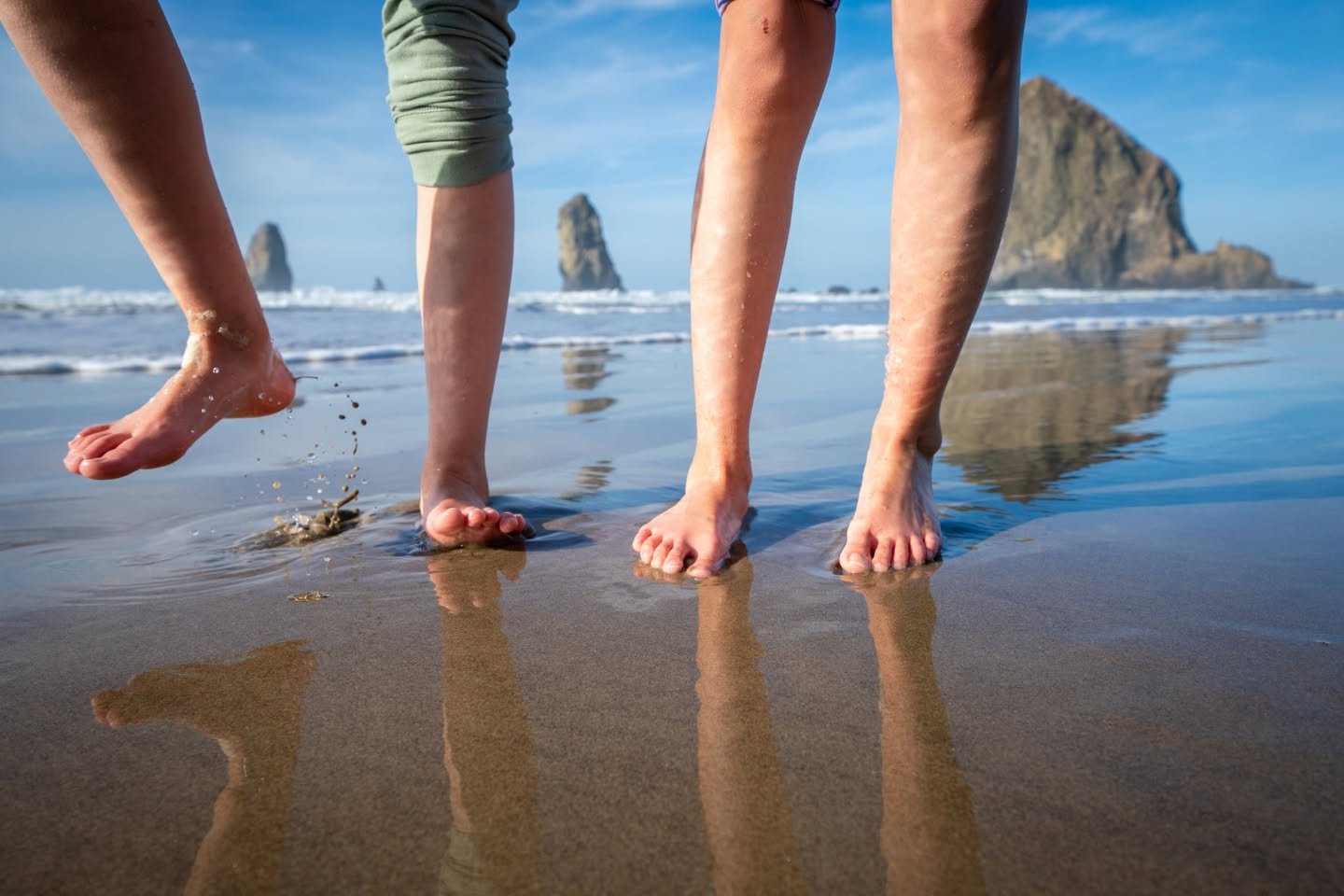 Barefoot individuals walking on a reflective wet sandy beach with rocky headlands and ocean waves in the distance under a blue sky.