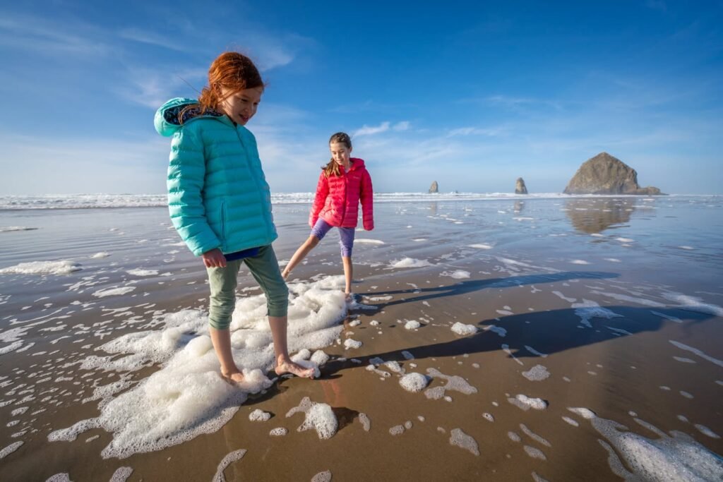 Two people walking barefoot on a wet, sandy beach with foam under a clear blue sky. A large sea stack is visible in the distance on the horizon.