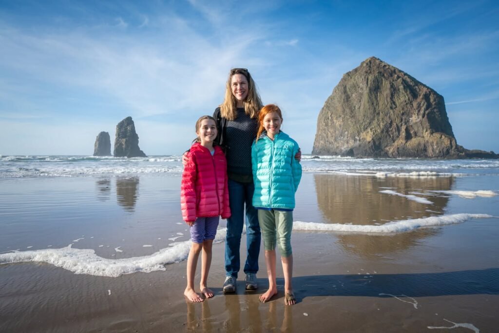 Three people pose on wet sand in front of Haystack Rock. The iconic sea stack and a distant rock formation rise from the Pacific Ocean under a blue Oregon sky.
