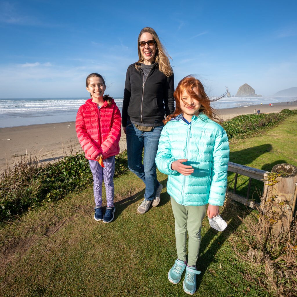 Three people walk along a grassy dune path beside the Pacific Ocean at Cannon Beach, Oregon, with Haystack Rock visible in the distance.