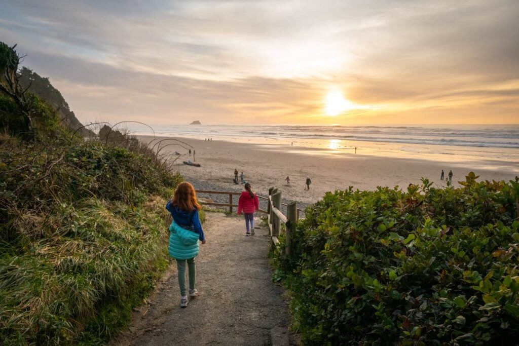 Two people walk down a sandy path toward a flat beach at sunset with the sun low over the ocean horizon.