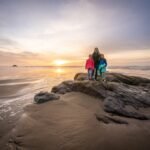 Three people standing on a rocky outcrop while the sun sets over the ocean, with soft golden light reflecting on the calm water.