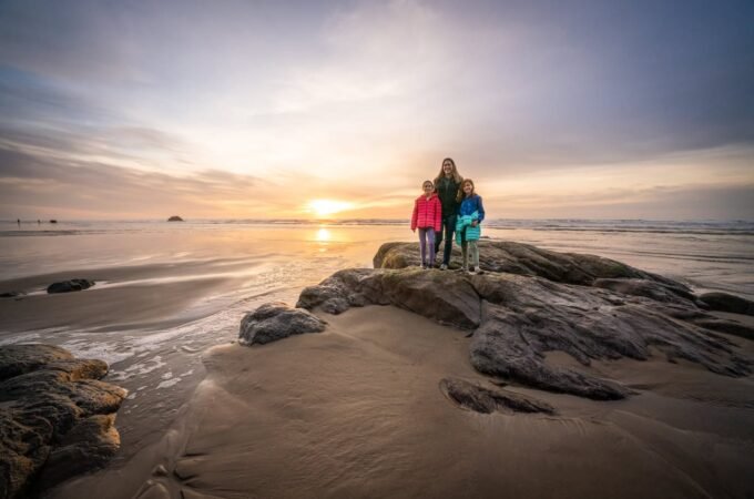 Three people standing on a rocky outcrop while the sun sets over the ocean, with soft golden light reflecting on the calm water.