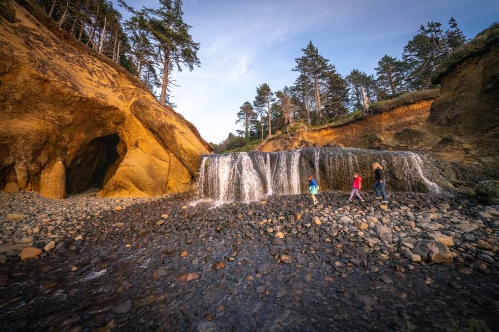 A wide shot of a wide, low waterfall running over dark basalt rocks in a forested canyon. Visitors explore the rocky terrain near the base of the falls at dusk.