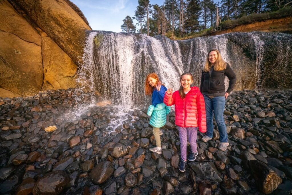 Three people standing on river rocks in front of a waterfall surrounded by trees.