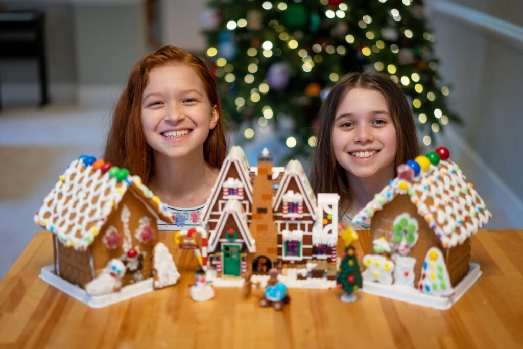 A panoramic view of a decorated gingerbread village with two cheerful individuals, with a detailed background featuring a Christmas tree. The display includes intricate icing work, miniature figures, and a colorful, cheerful setting.