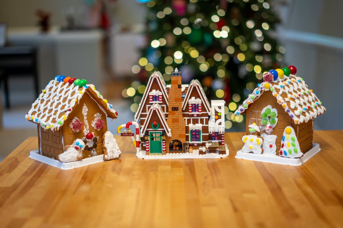 Gingerbread Houses Three elaborately decorated gingerbread houses on a wooden surface with a Christmas tree in the background.