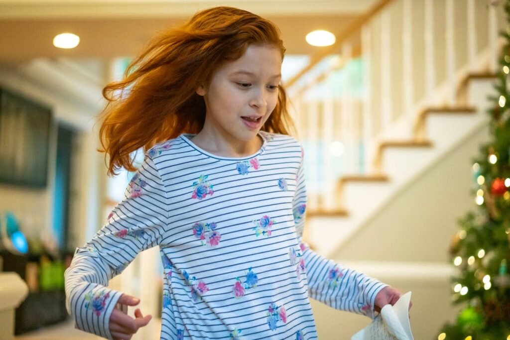 A child in festive pajamas moving near a decorated staircase in a cozy living room.