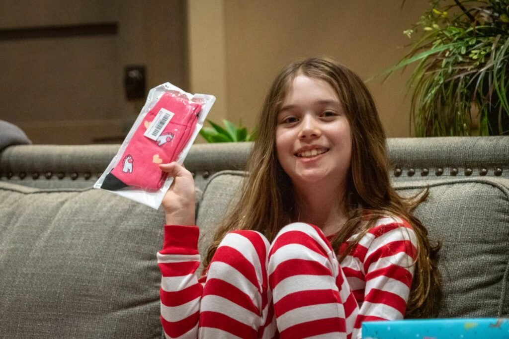 A young person in a red and white striped top holding a pink item on a gray couch. Natural indoor lighting highlights the green plants in the background.