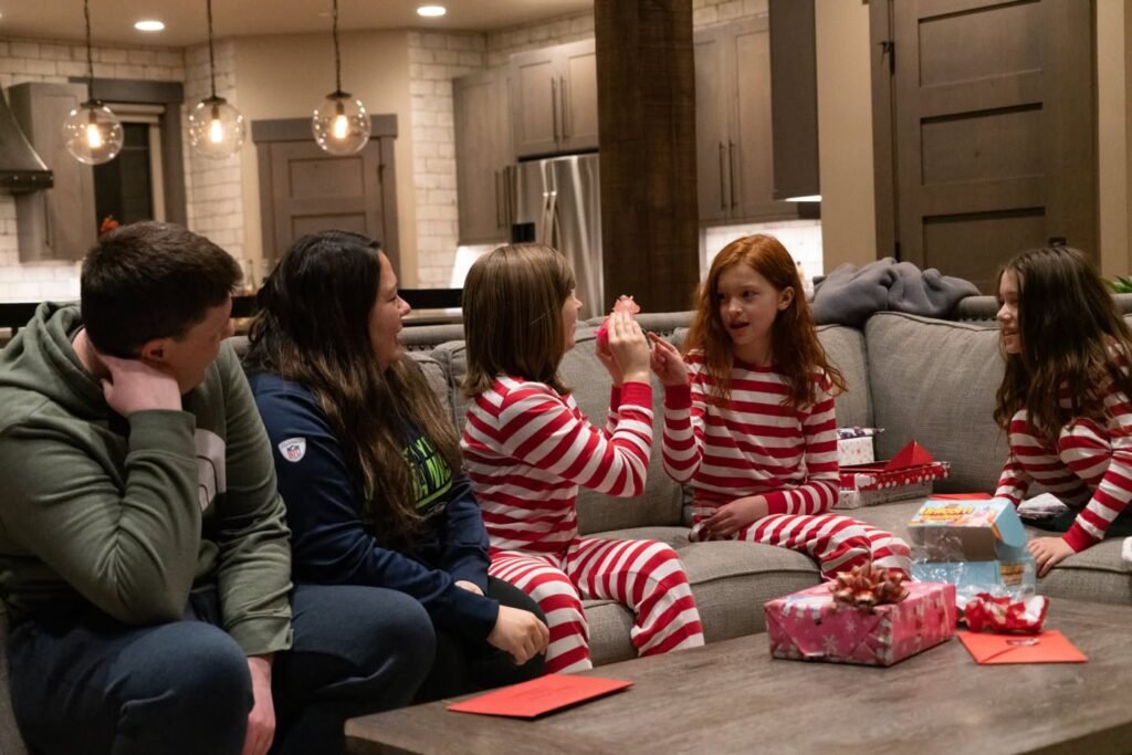 A family gathered on the living room couch wearing matching red and white striped pajamas, surrounded by wrapped Christmas gifts and orange papers.
