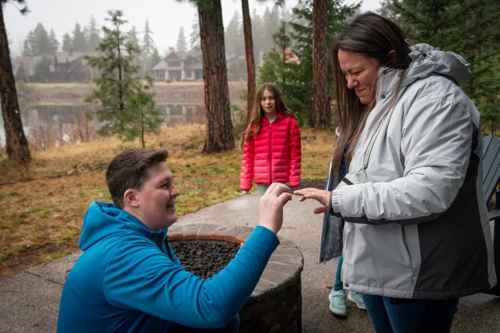 Family sharing a moment around a fire pit by a misty lake. Tall evergreens and a distant cabin complete the scenic outdoor setting.