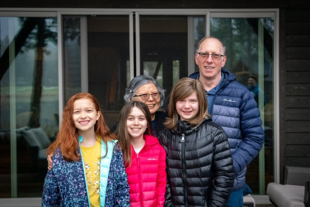 A happy group of people wearing winter jackets poses for a photo outside a modern wooden cabin with large glass windows. The background features reflections of trees, suggesting a natural, outdoor setting.