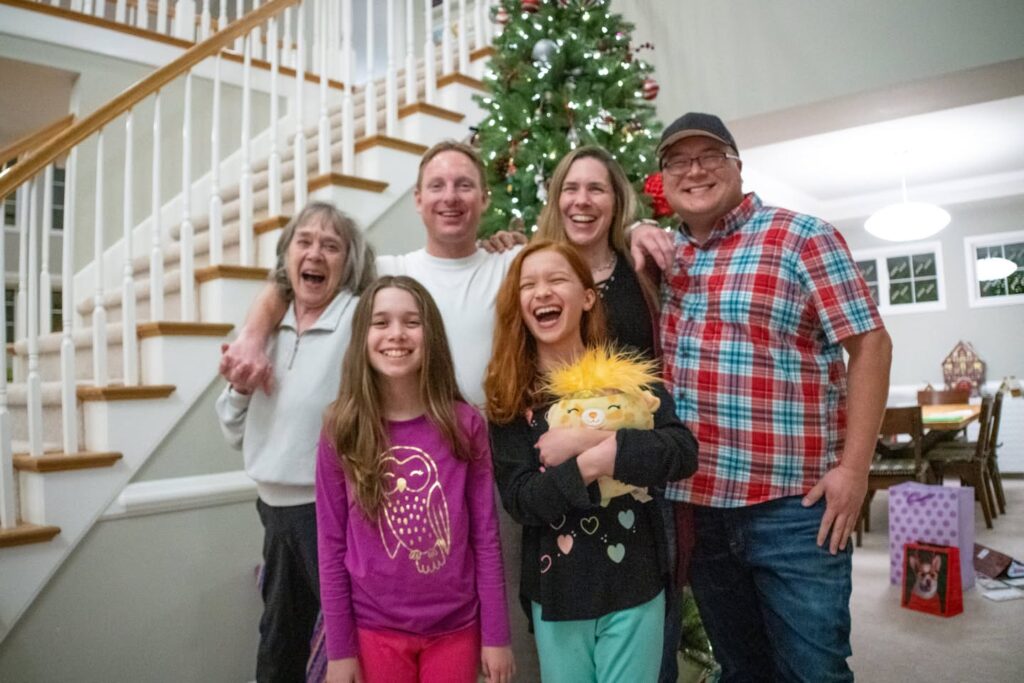 A happy family gathers around a decorated Christmas tree in a bright living room. The festive scene includes gifts, holiday cheer, and warm smiles from all ages.