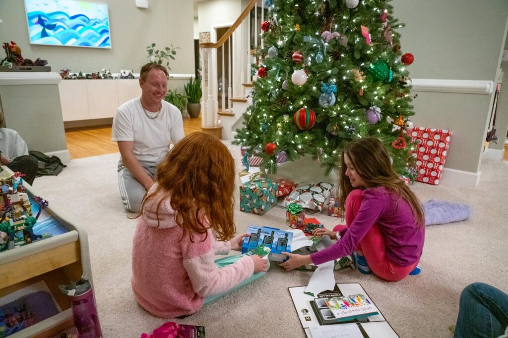 A family gathered on the carpet in front of a decorated Christmas tree playing with colorful toys on a bright winter morning.