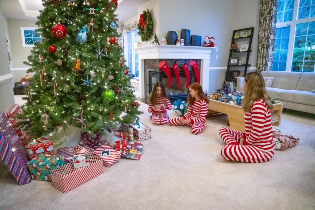 A cozy living room decorated for the holidays with a large tree, gifts, red stockings on the fireplace, and individuals in matching pajamas gathering around the tree.