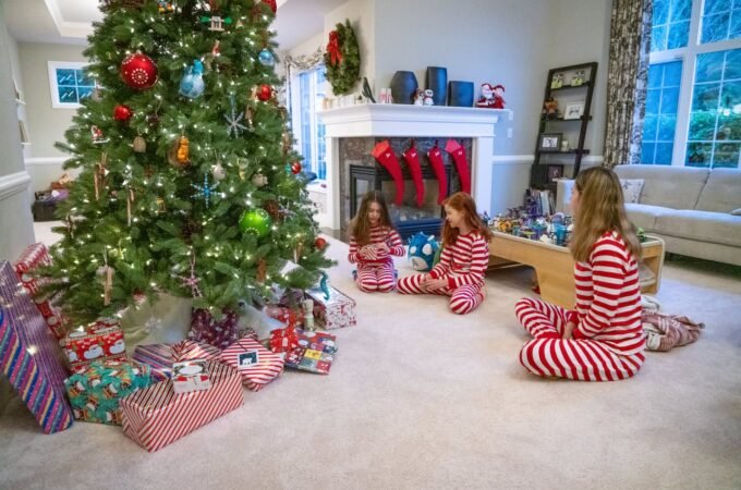 A cozy living room decorated for the holidays with a large tree, gifts, red stockings on the fireplace, and individuals in matching pajamas gathering around the tree.