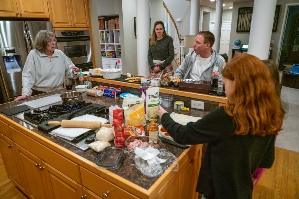 Several people gathered around a kitchen island preparing food. Various baking ingredients and a rolling pin are visible on the countertop.