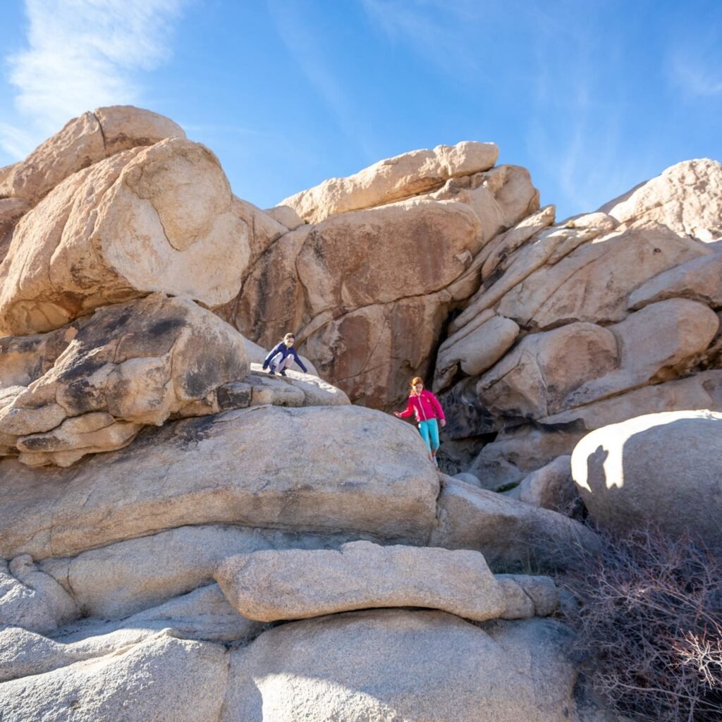 Hikers navigating a granite boulder field under a bright blue sky. The rugged terrain features smoothed rock formations typical of a desert landscape.