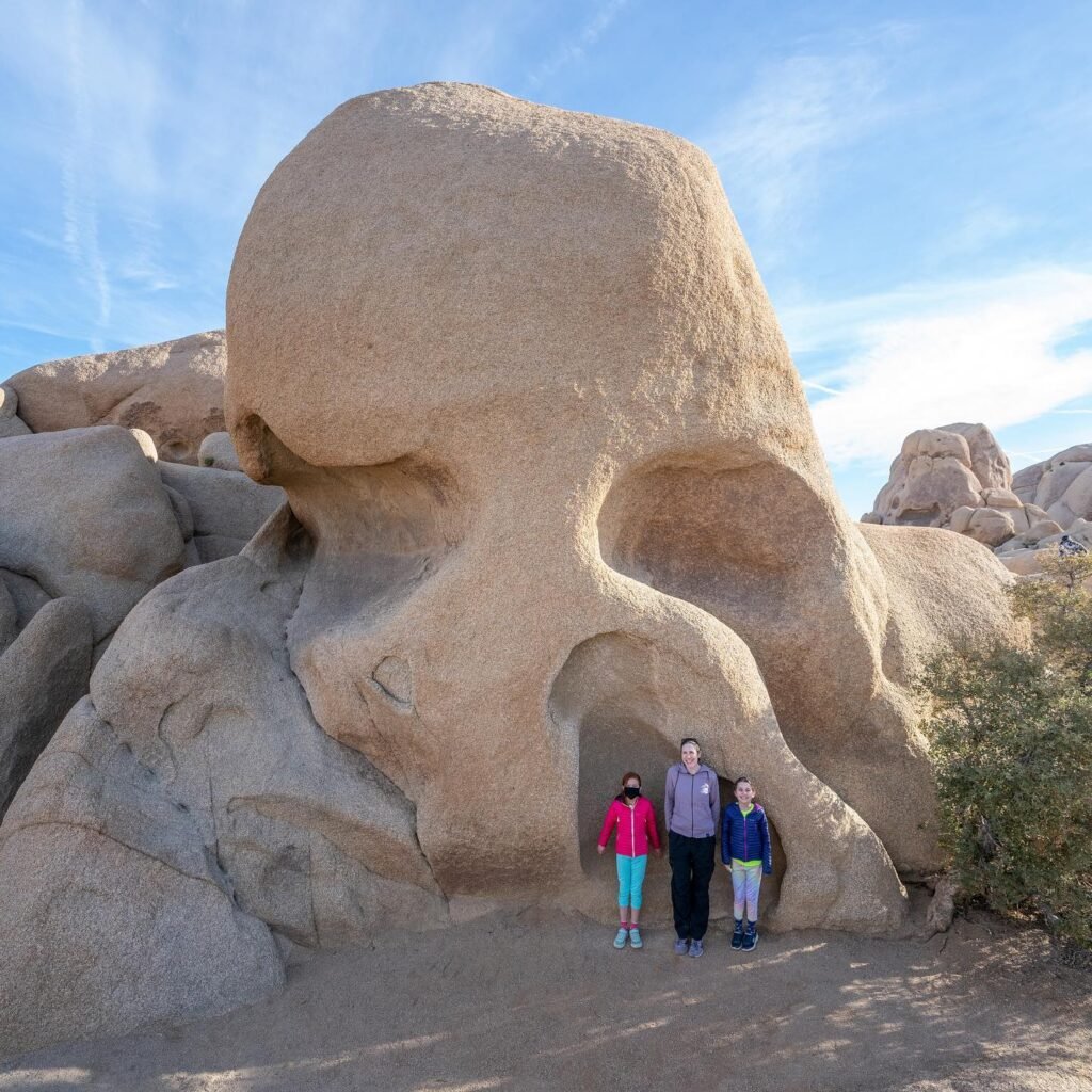 A large natural rock formation shaped like a human skull with three people standing in front of it under a clear blue sky.
