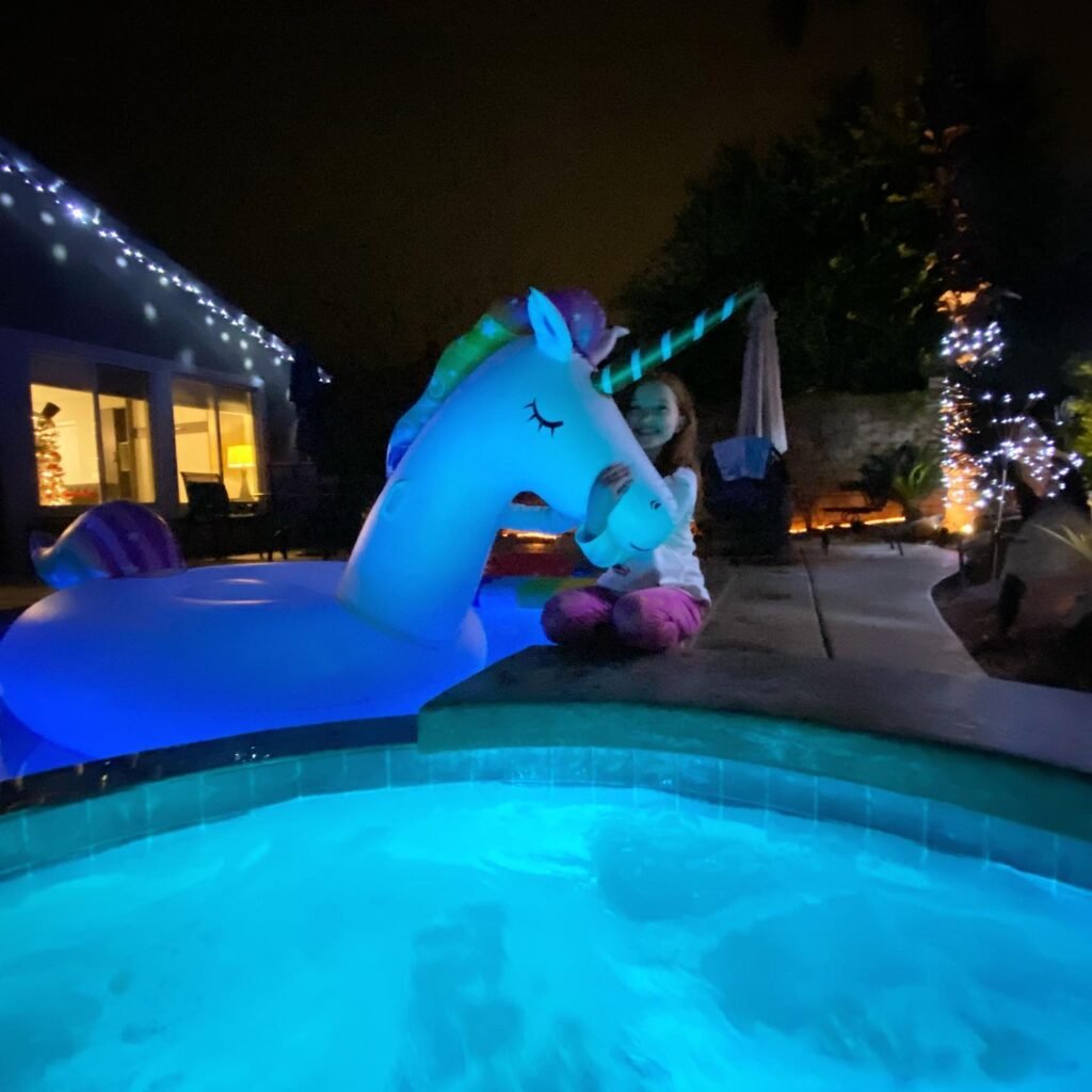 A glowing blue unicorn pool float at the edge of a pool in a backyard with string lights.
