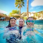 People wearing goggles in a clear blue pool with palm trees and desert landscaping in the background.