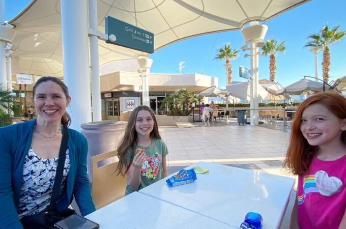 People enjoying a sunny day at an outdoor plaza with palm trees and modern architecture.