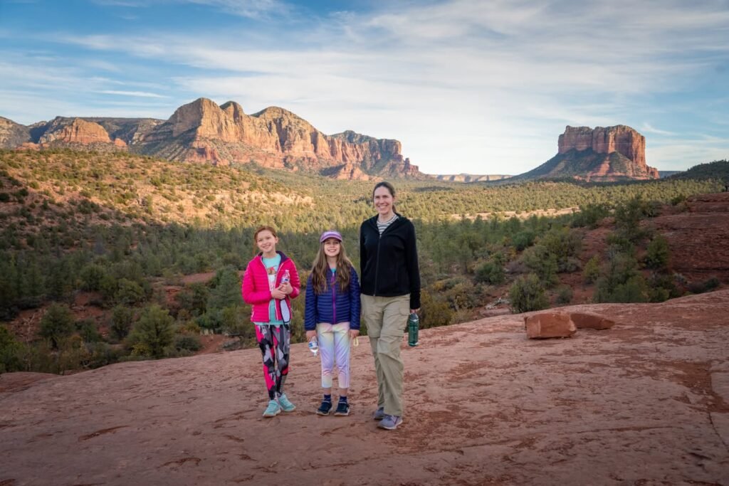 Three family members enjoy a scenic hiking spot with red rock formations and desert vegetation in the background.