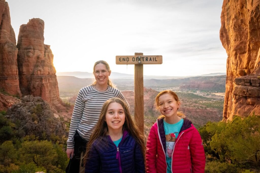 A family poses at the end of a trail marker in a rugged red rock canyon with a scenic overlook.