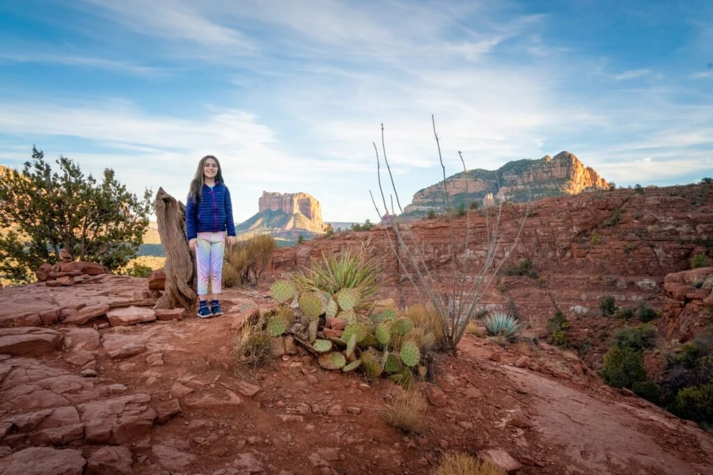 A person in a blue jacket stands by a rocky red desert landscape with prickly pear cacti and a distant mesa.