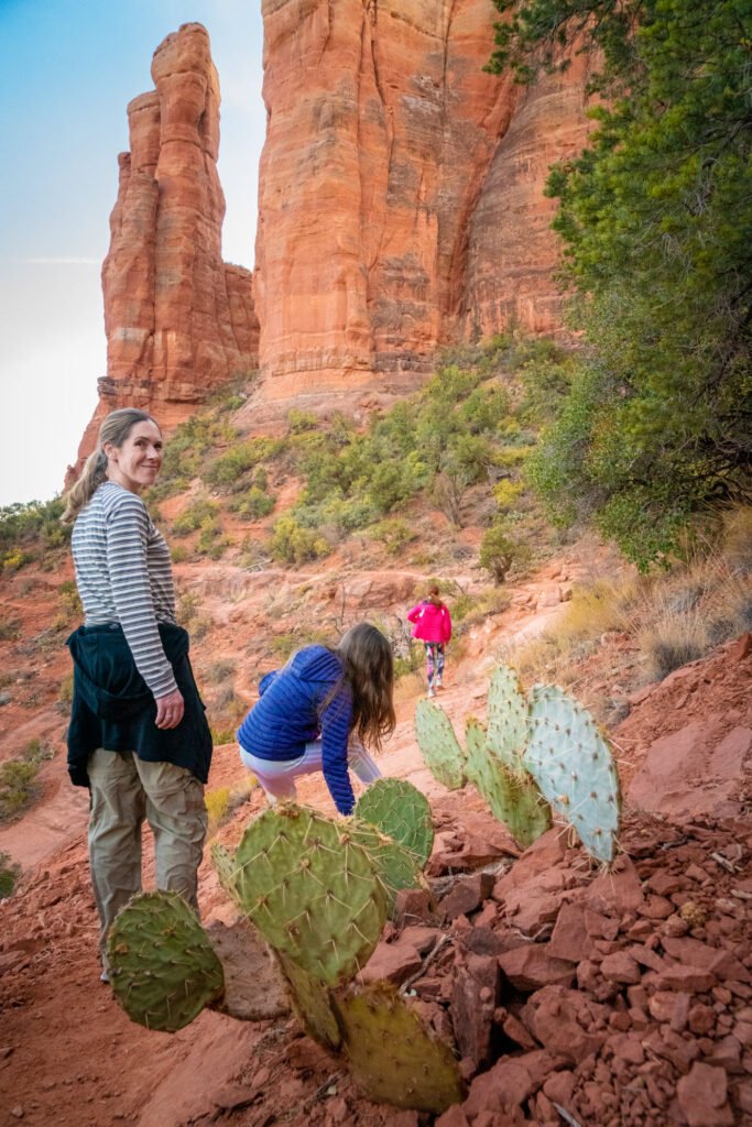 Hikers explore a rugged desert trail featuring striking red rock formations and prickly pear cacti. The scene captures the beauty of the Southwest landscape under a clear blue sky.