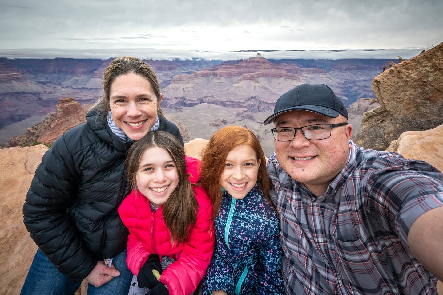 The Grand Canyon – Ooh Aah Point A happy family poses for a selfie at a scenic canyon overlook. The backdrop features a vast desert landscape under a cloudy sky.