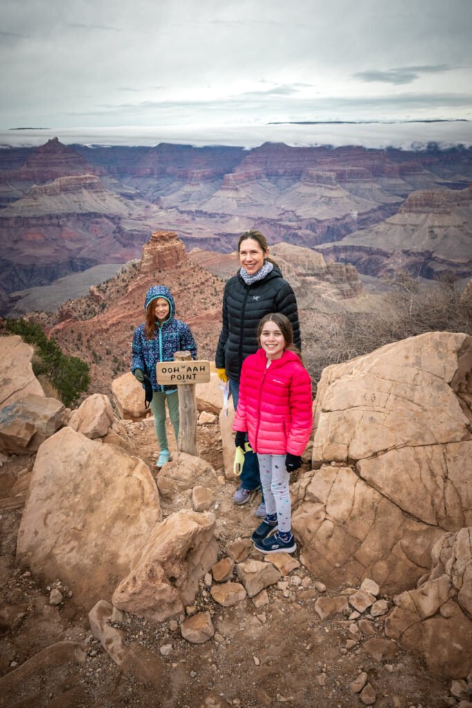 A family poses on a rocky overlook with sweeping views of the canyon below under a cloudy sky. The colorful jackets stand out against the rugged natural landscape.