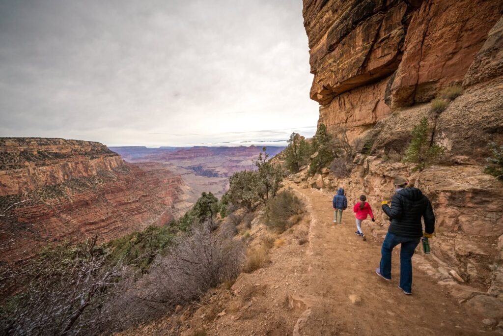 A group of hikers walks along a dirt path on a sandstone cliff edge overlooking a vast desert canyon under a cloudy sky.