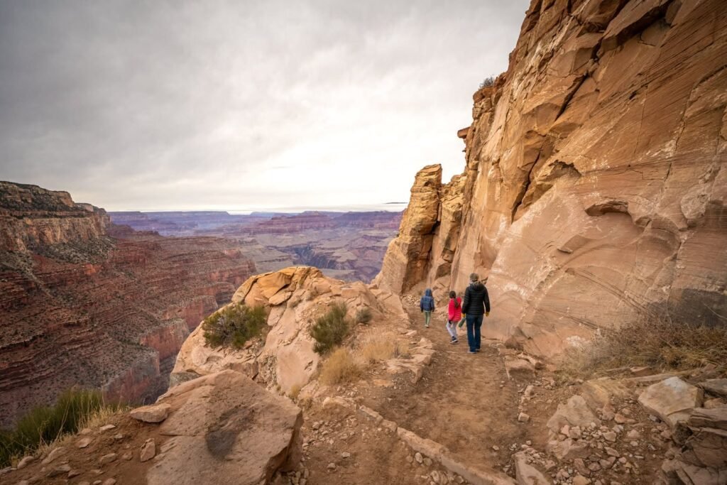 Hikers walk along a rugged trail beside towering sandstone cliffs under an overcast sky in Arches National Park.