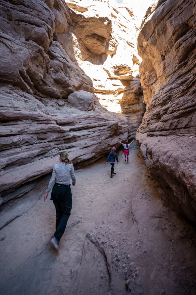 Hikers walking through a narrow slot canyon with tall, textured sandstone walls and a sandy floor.
