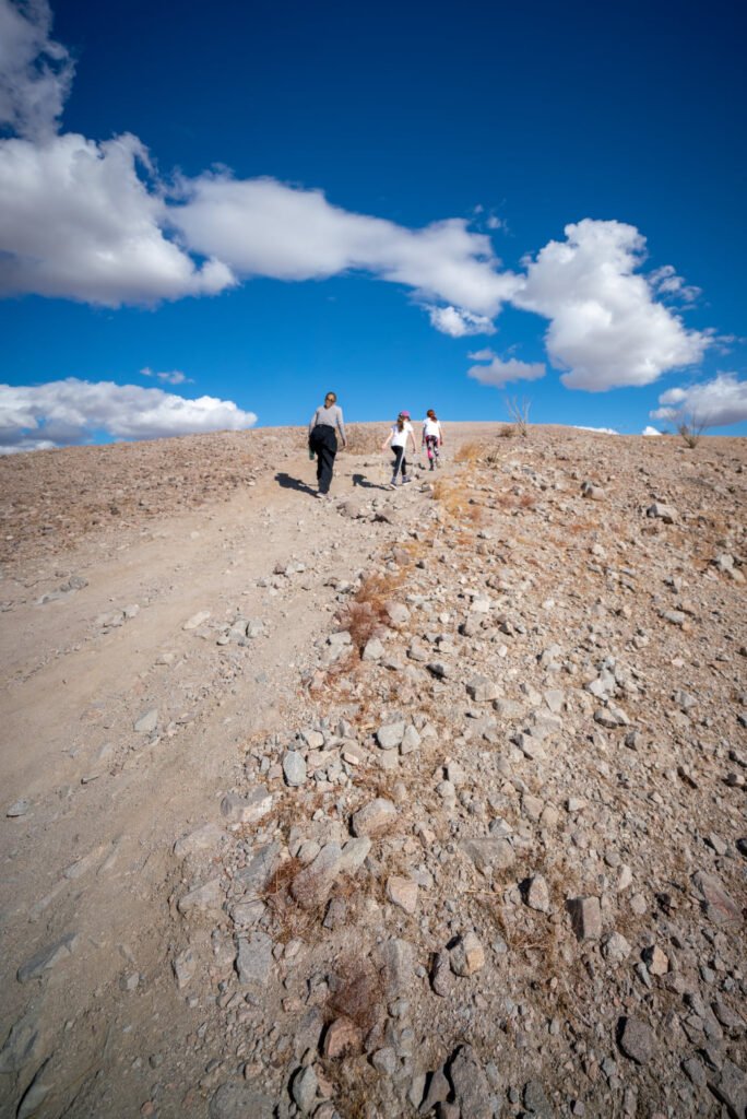 A group of people hiking on a rocky mountain path under a bright blue sky with white clouds.